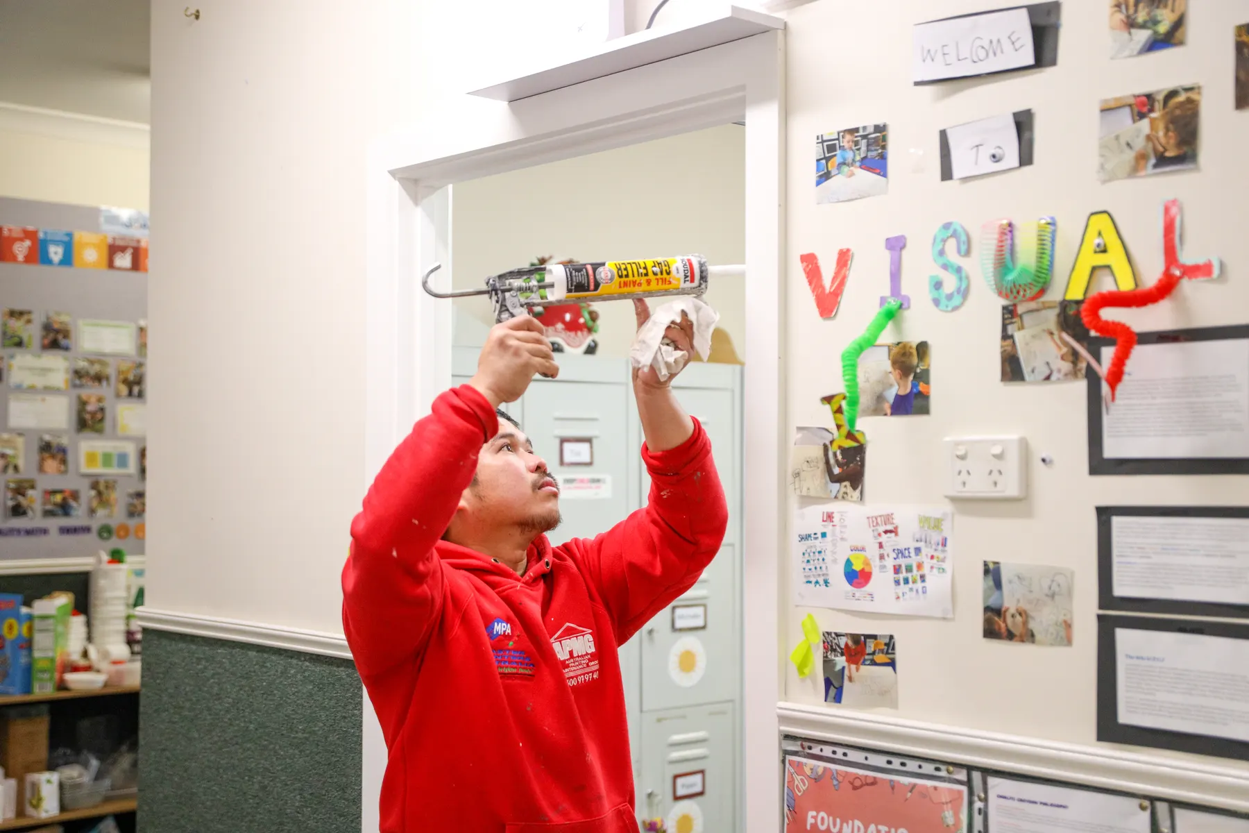A painter in company uniform running gap filler along the top of a door frame, children's artwork on the wall beside it