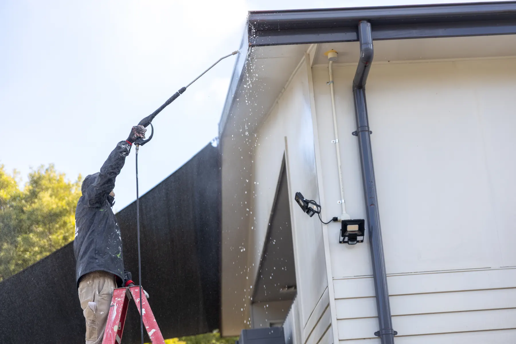 A painter on a stepladder pressure washing the fascia and gutter line of a weatherboard building, spray falling past a downpipe