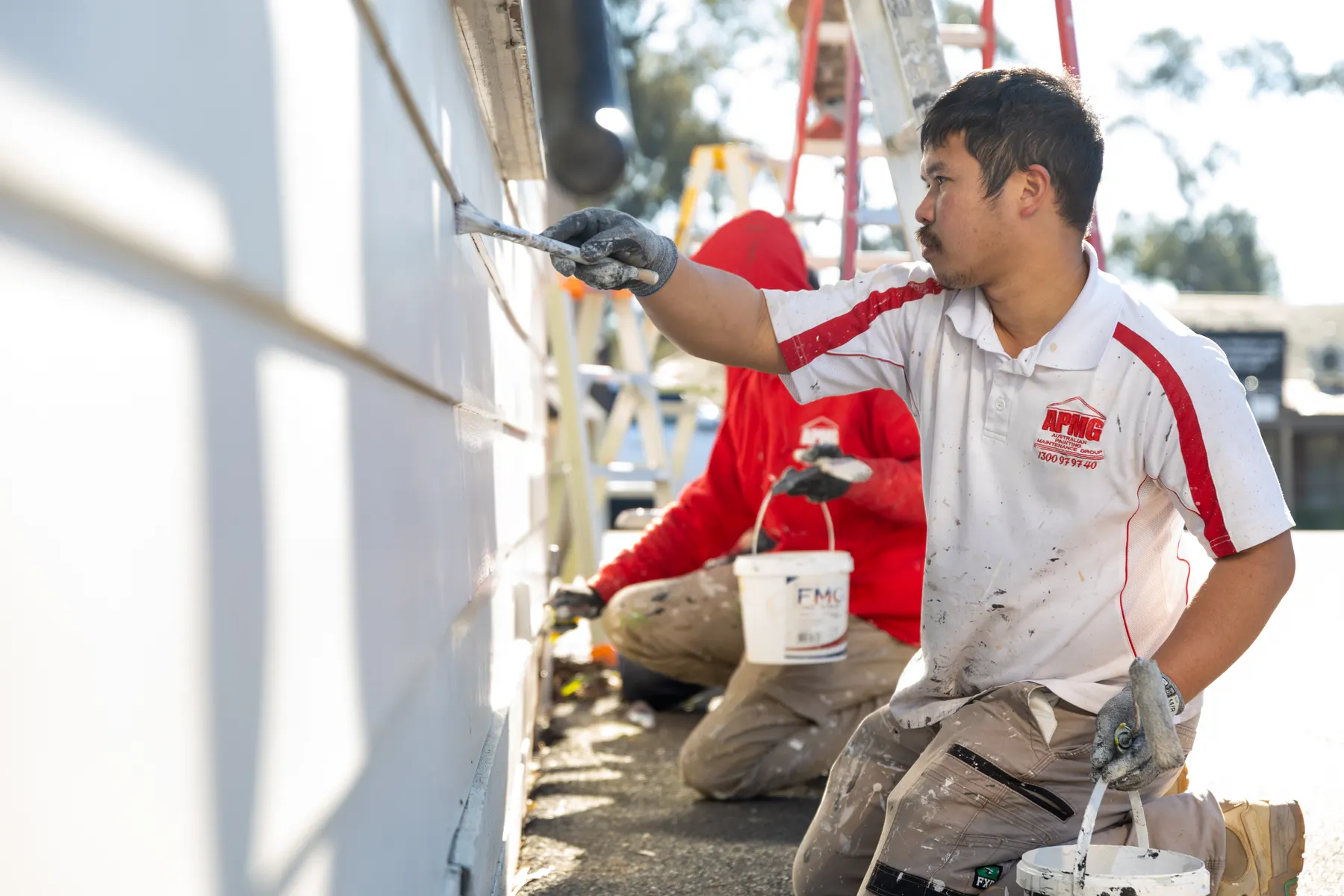 A painter in company uniform brushing along a weatherboard line, a second painter crouched behind him with a pail, stepladders beyond