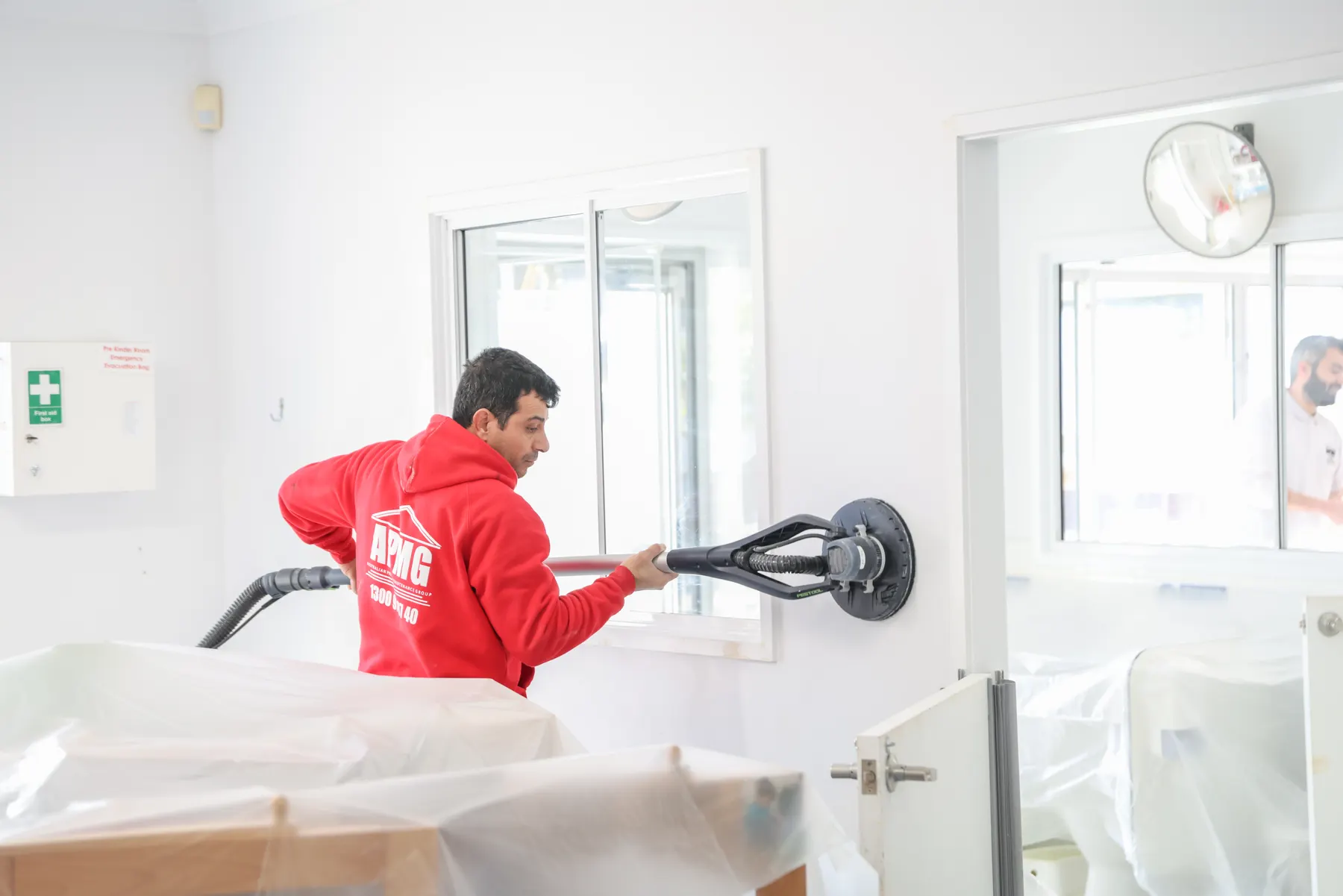A painter in company uniform sanding a wall with a vacuum-fed pole sander, room furniture under plastic sheeting in front of him