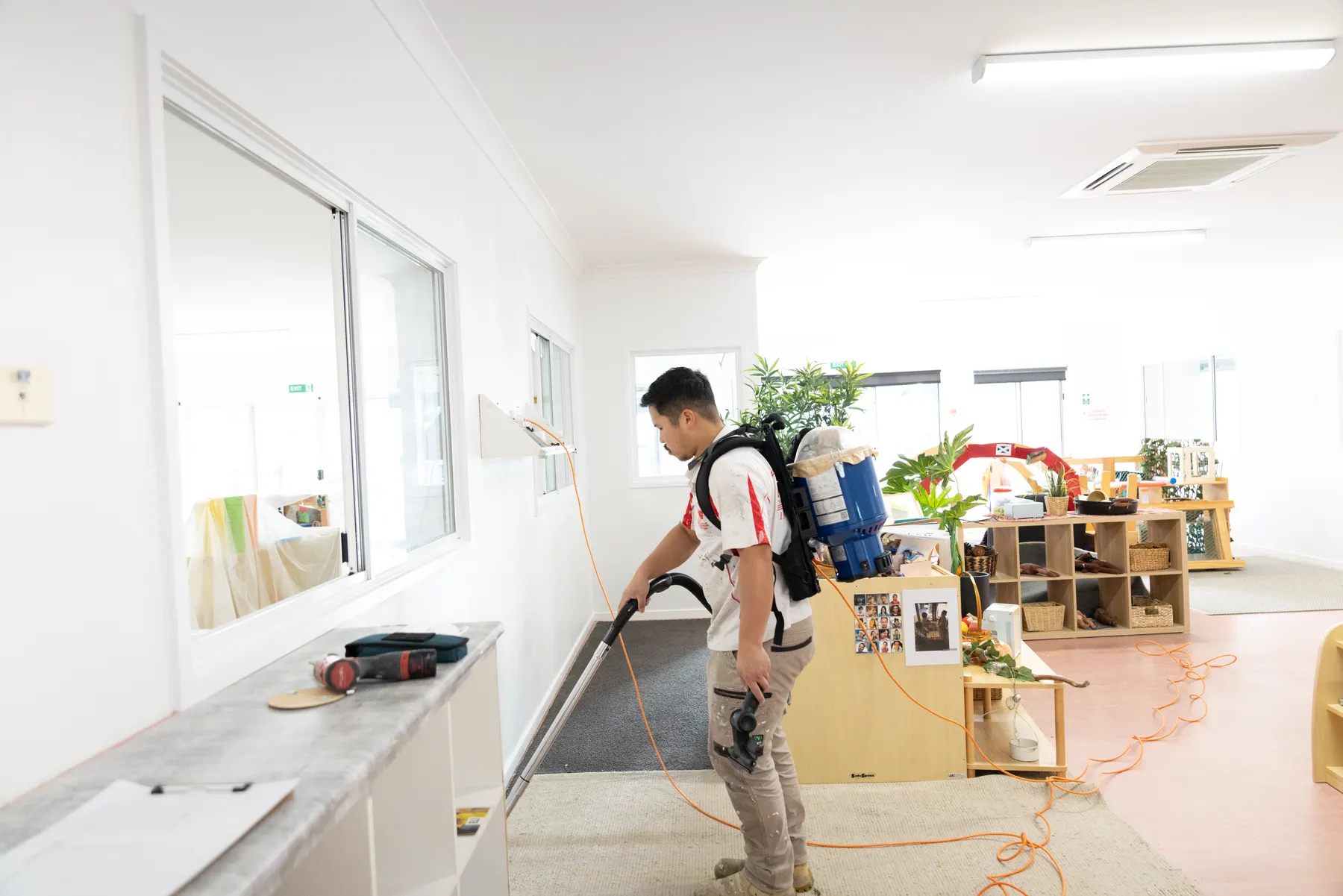 A crew member running a backpack vacuum along the edge of a childcare room carpet, play furniture and shelving in place behind