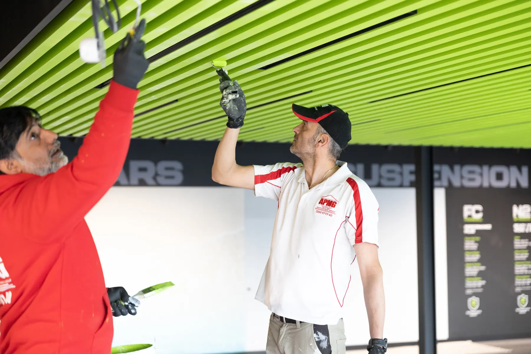 Two painters in company uniform rolling a lime-green slatted ceiling with small rollers, showroom wall signage behind