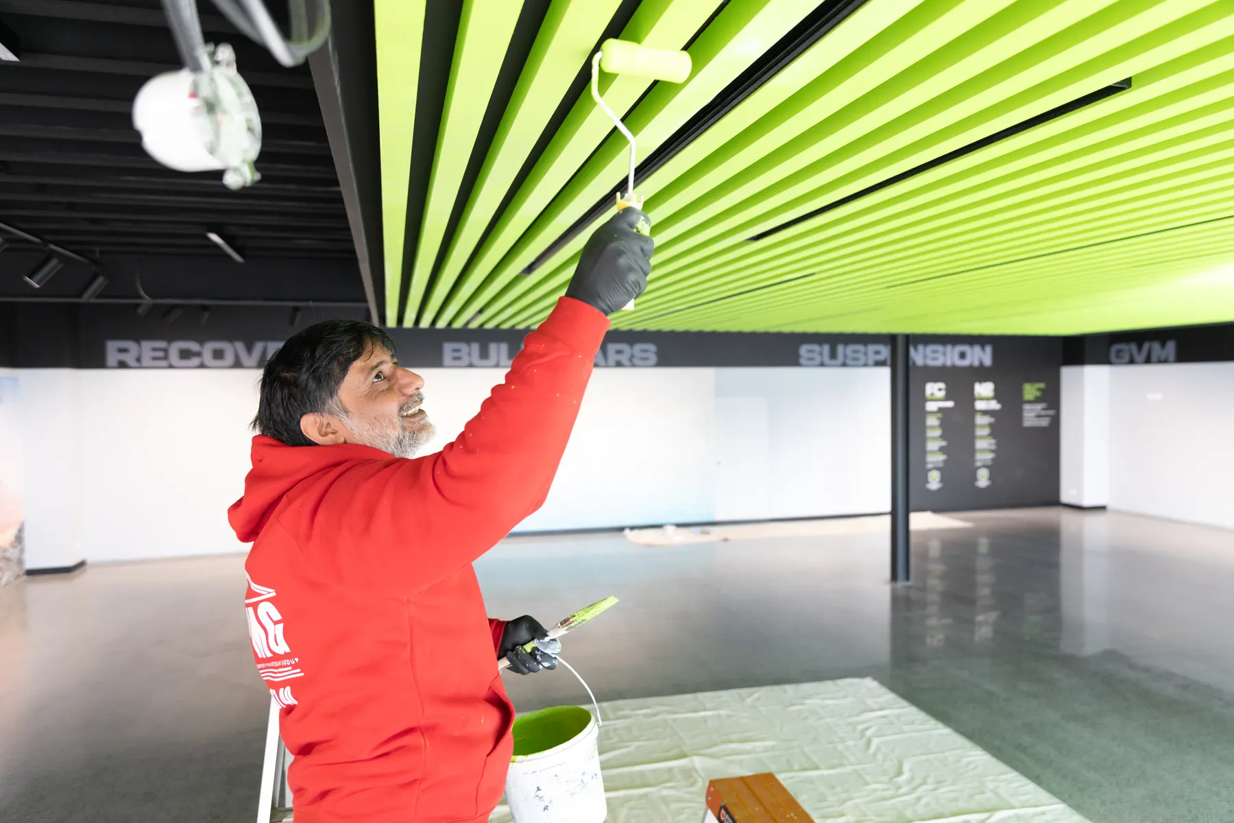 A painter in company uniform rolling the underside of a lime-green slatted ceiling, drop sheets on the polished showroom floor