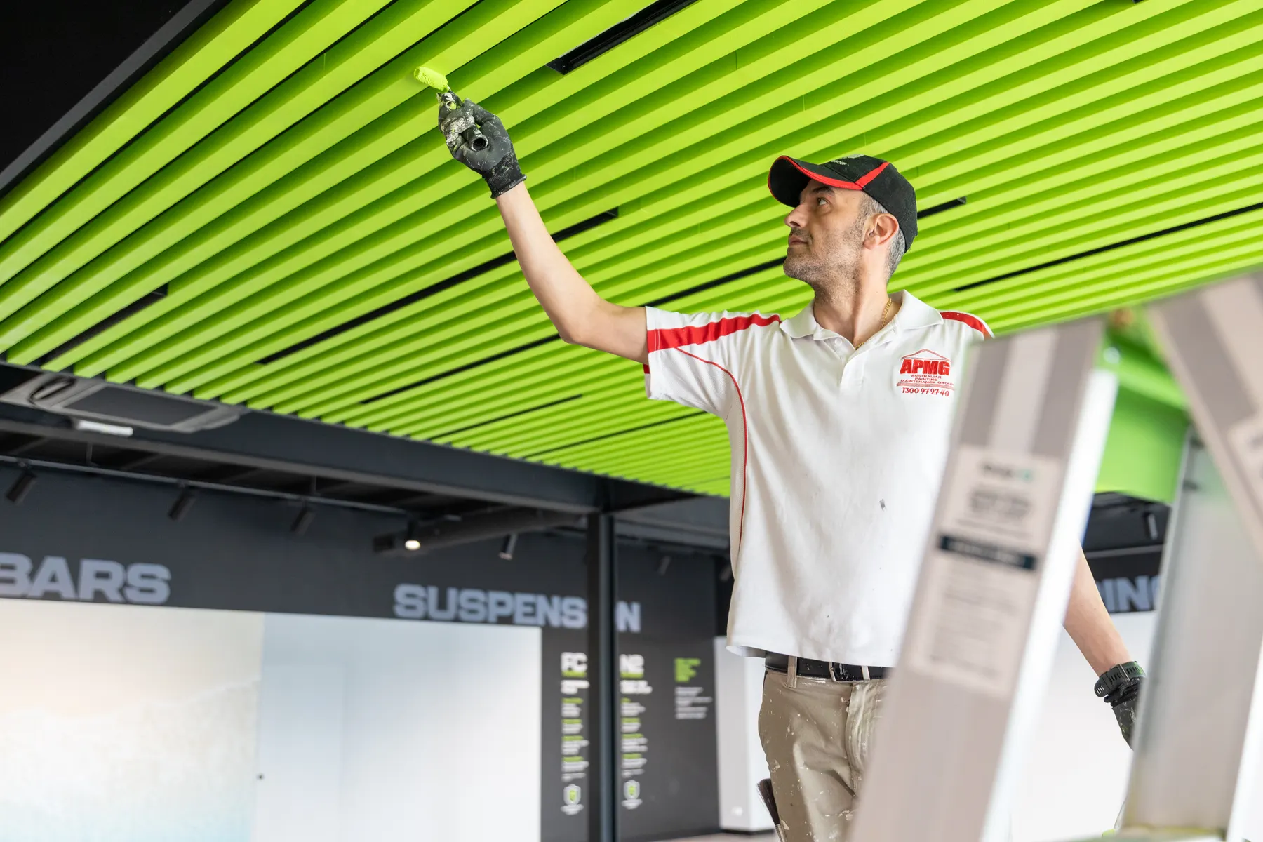 A painter in company uniform reaching up with a small roller to a lime-green slatted ceiling, a stepladder in the foreground