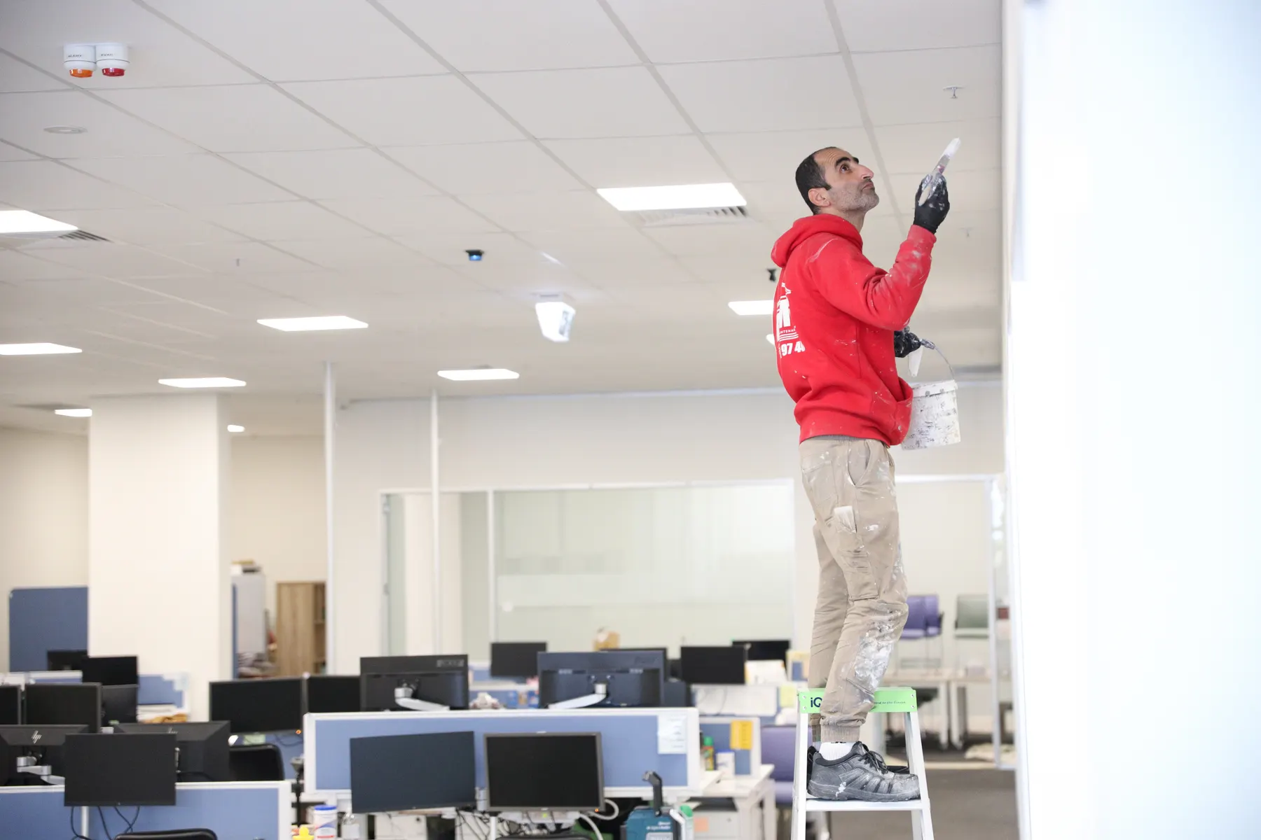 A painter in company uniform on a stepladder cutting in high on a wall above an open-plan office of workstations and monitors