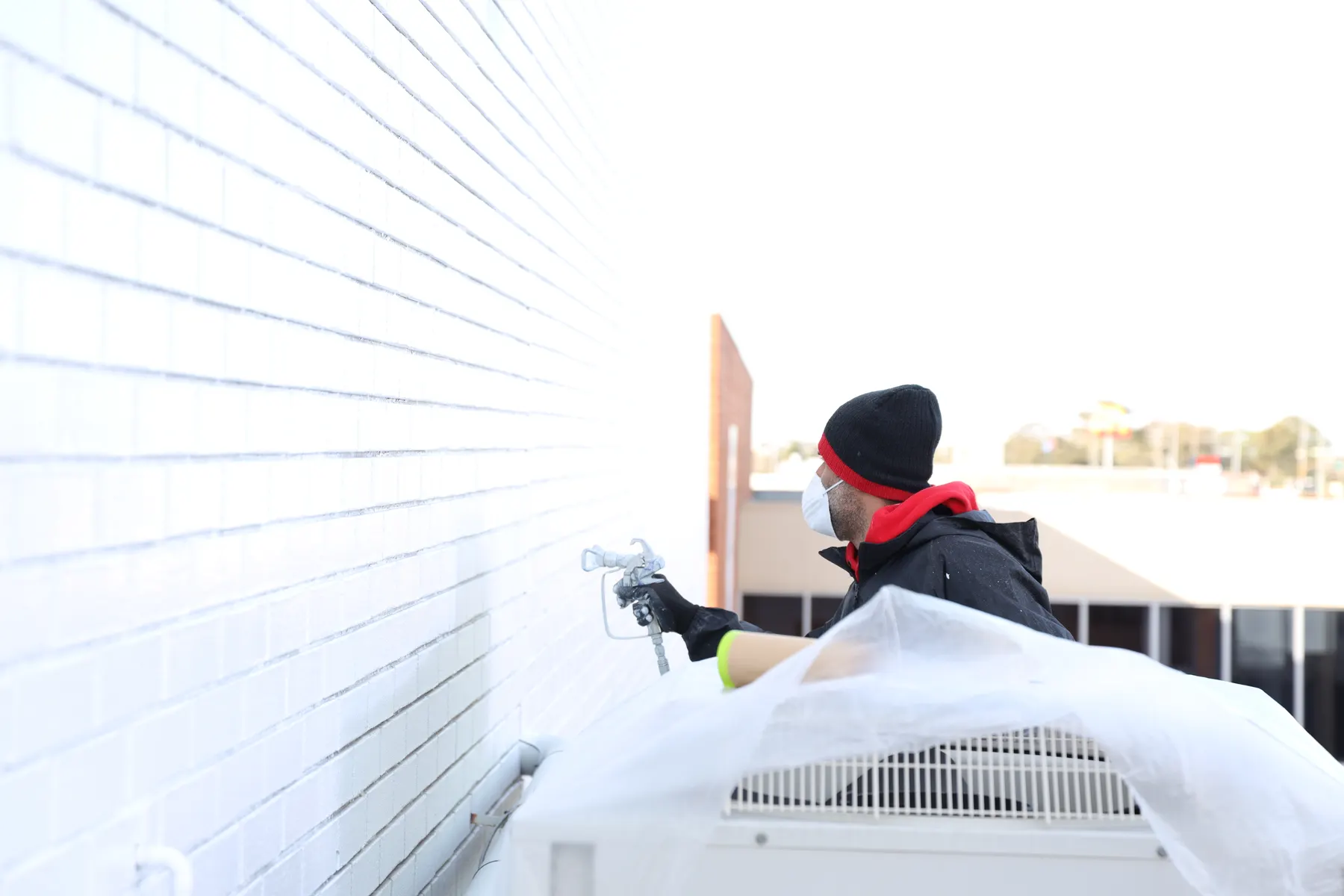 A painter in a dust mask spraying a white block wall beside an air conditioning unit wrapped in plastic sheeting