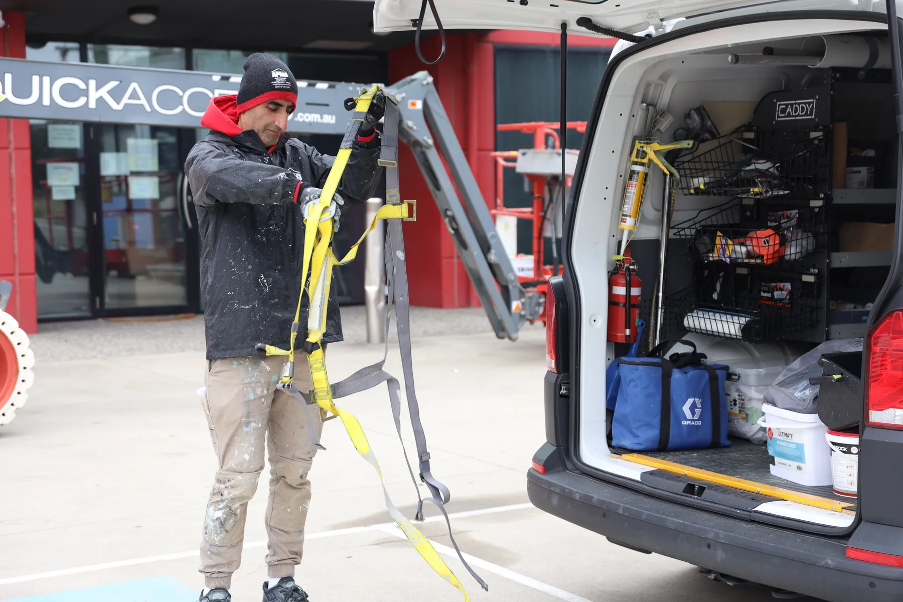 A painter in company uniform sorting a yellow safety harness at the back of a work van, a boom lift parked behind