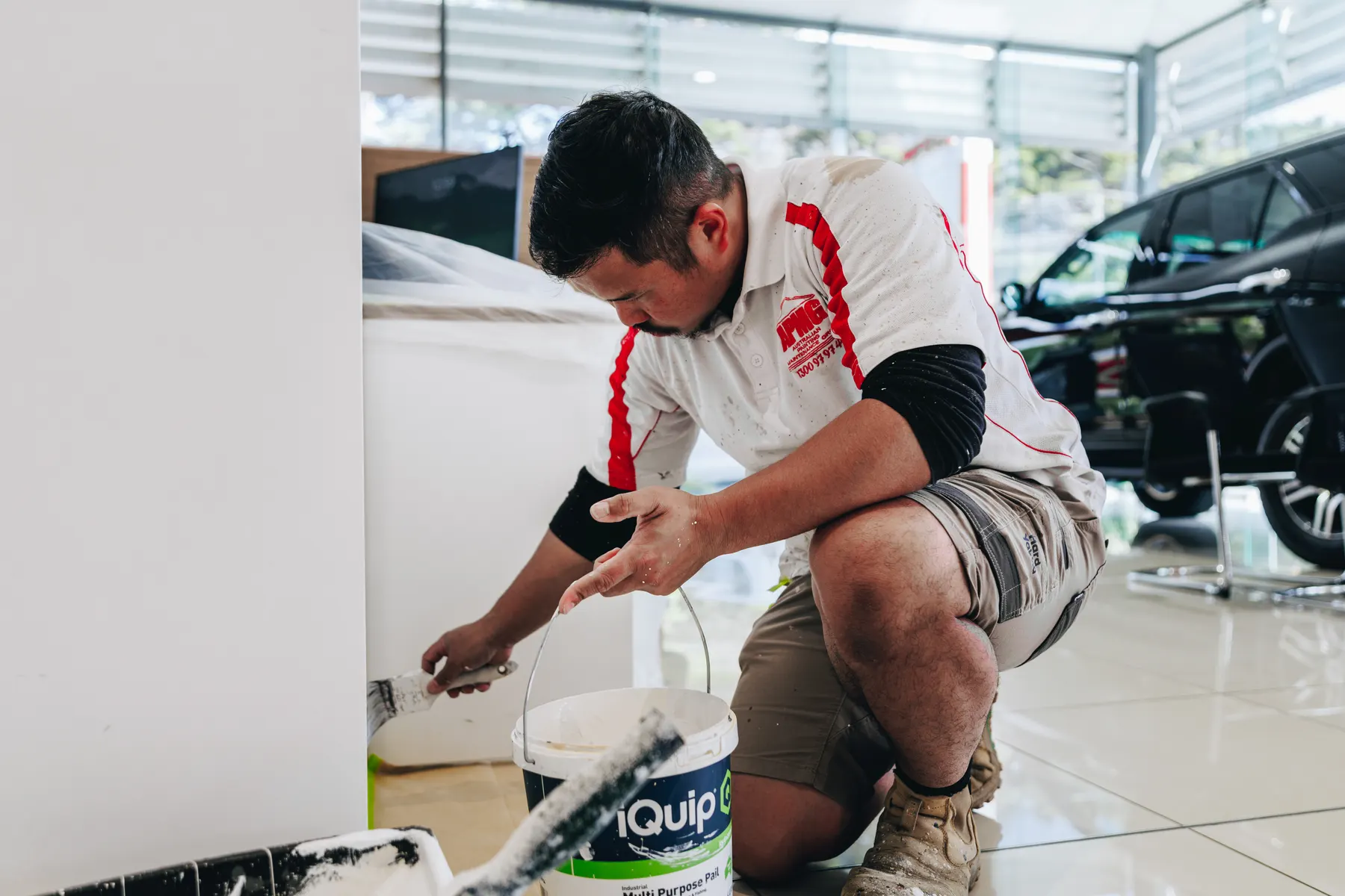 A painter in company uniform kneeling to cut in the base of a showroom wall with a brush beside a paint pail