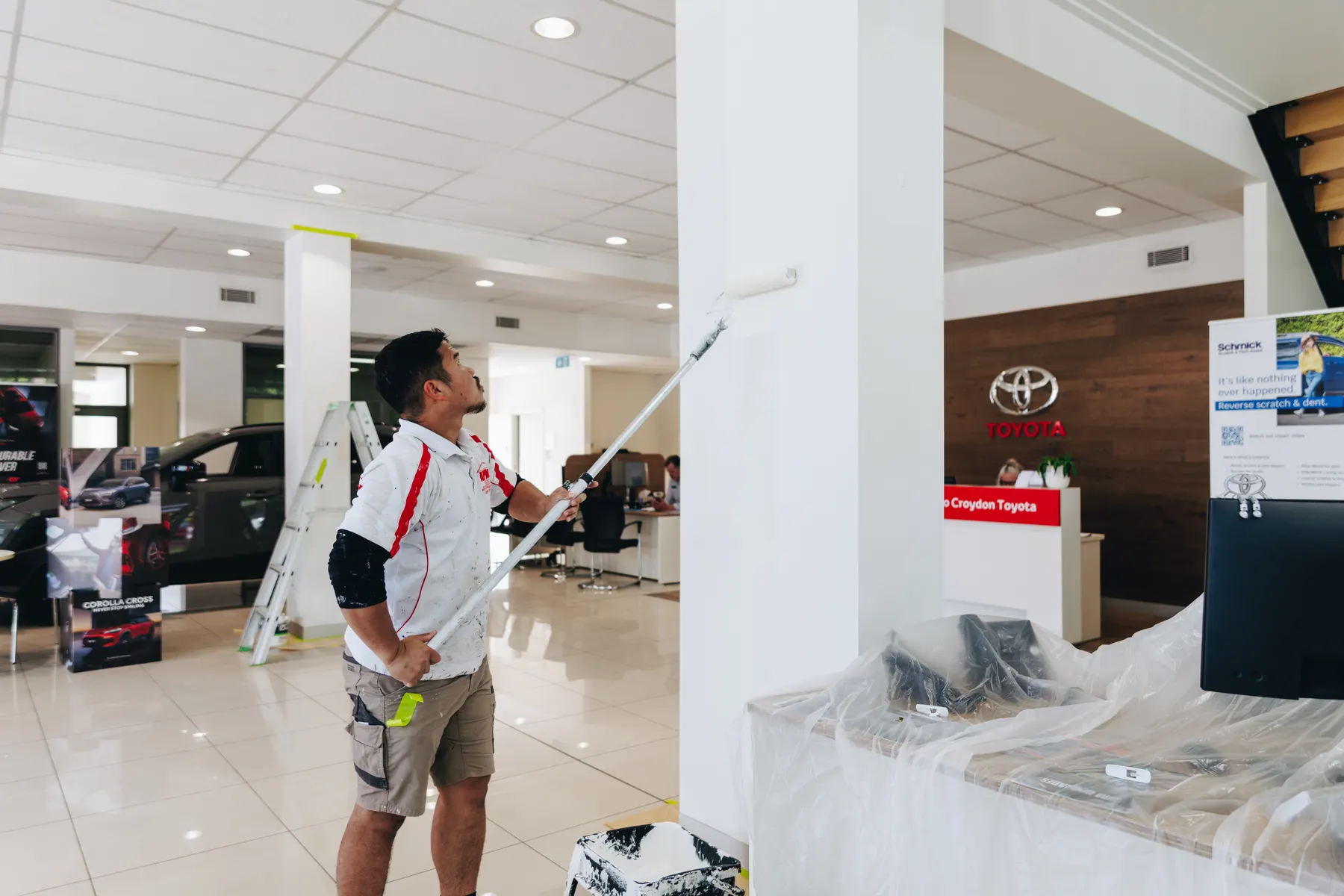 A painter rolling a column in the showroom with an extension-pole roller, stepladder and plastic-sheeted desk behind