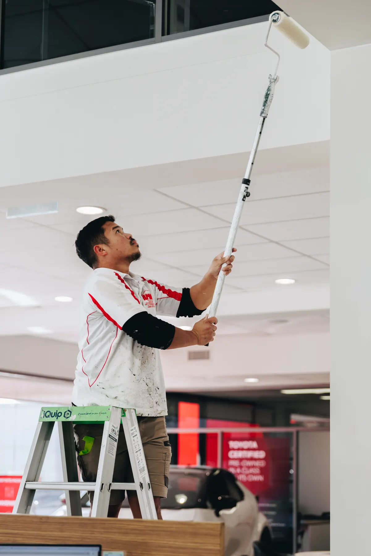 A painter on a stepladder running an extension-pole roller across a bulkhead above the showroom floor