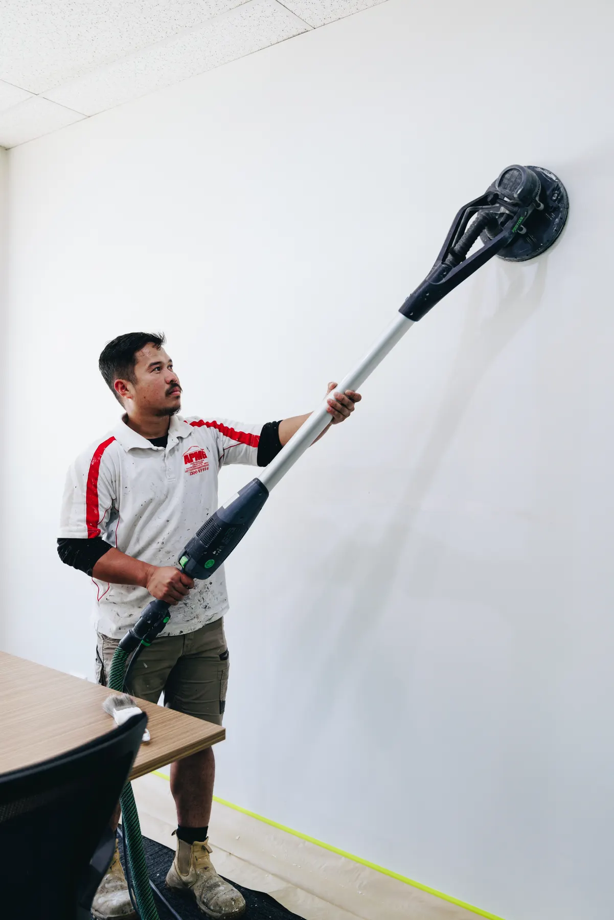 A painter sanding an office wall with a vacuum-fed pole sander, masking tape at the wall base and floor protection down