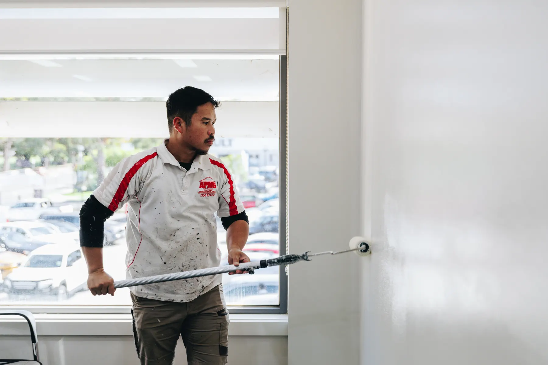 A painter in company uniform working an extension-pole roller across an interior wall, window and carpark behind