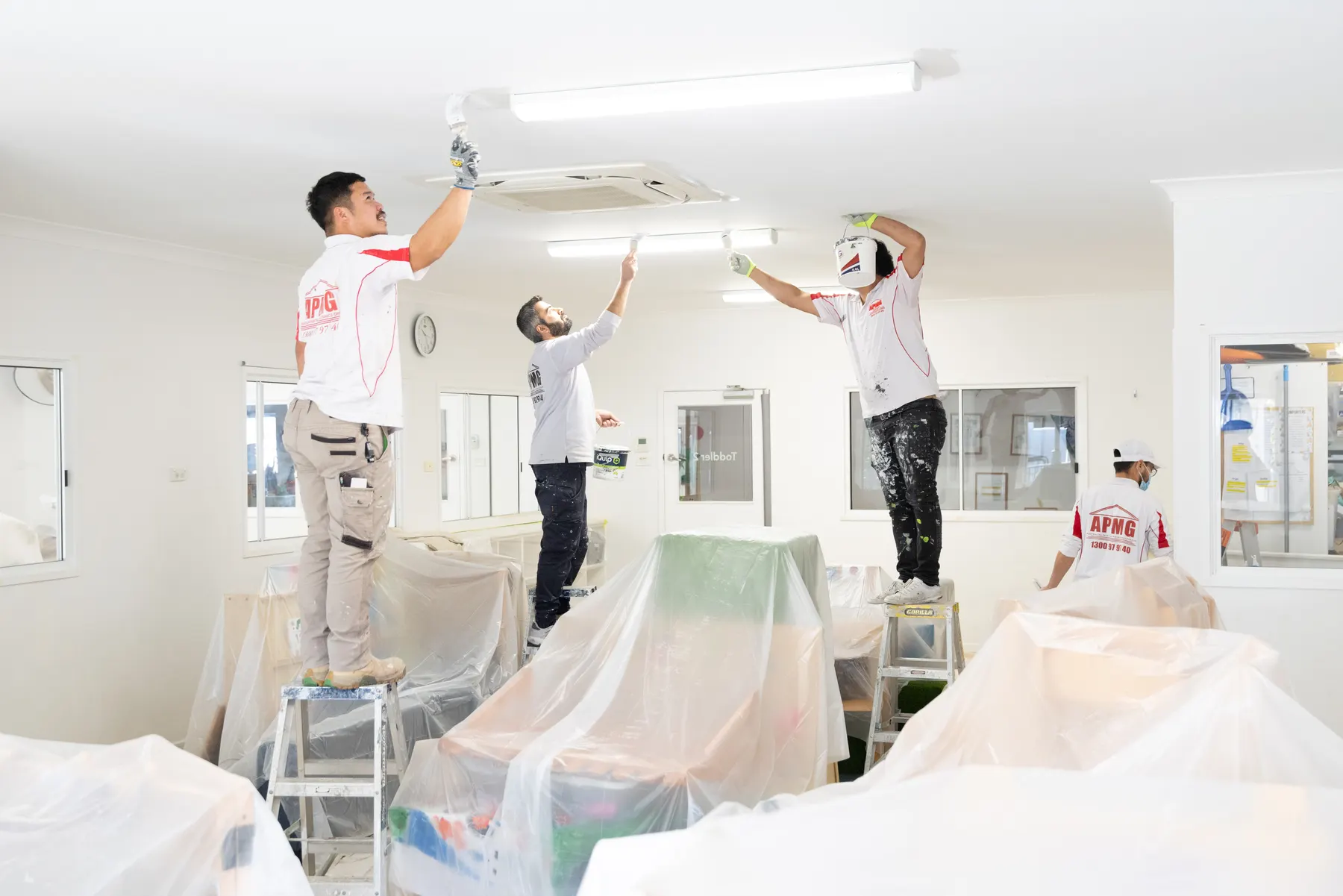 Three painters cutting in a childcare room ceiling, two on stepladders, while a fourth works the plastic sheeting over the furniture
