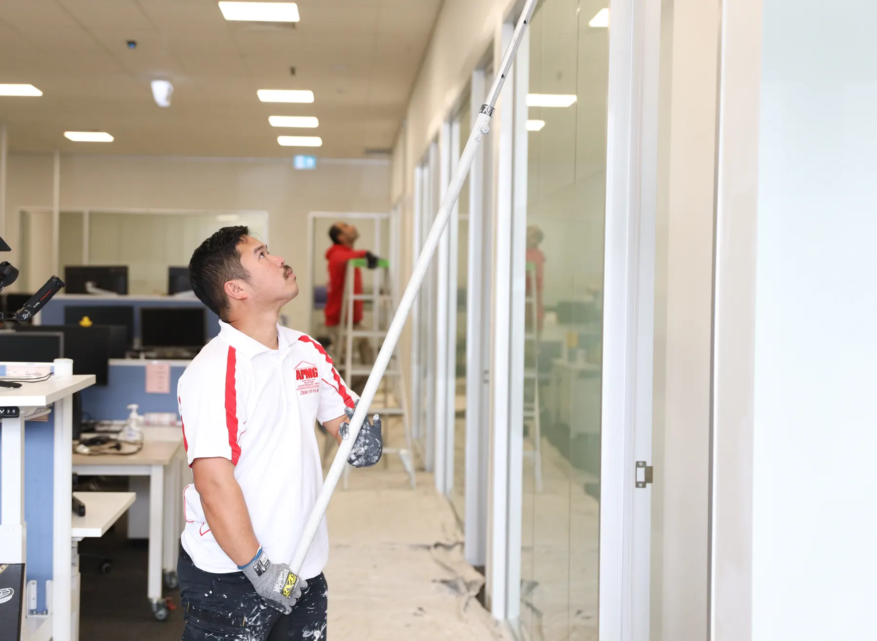 A painter in company uniform running an extension-pole roller up a wall beside glass offices, drop sheets down and a second painter behind