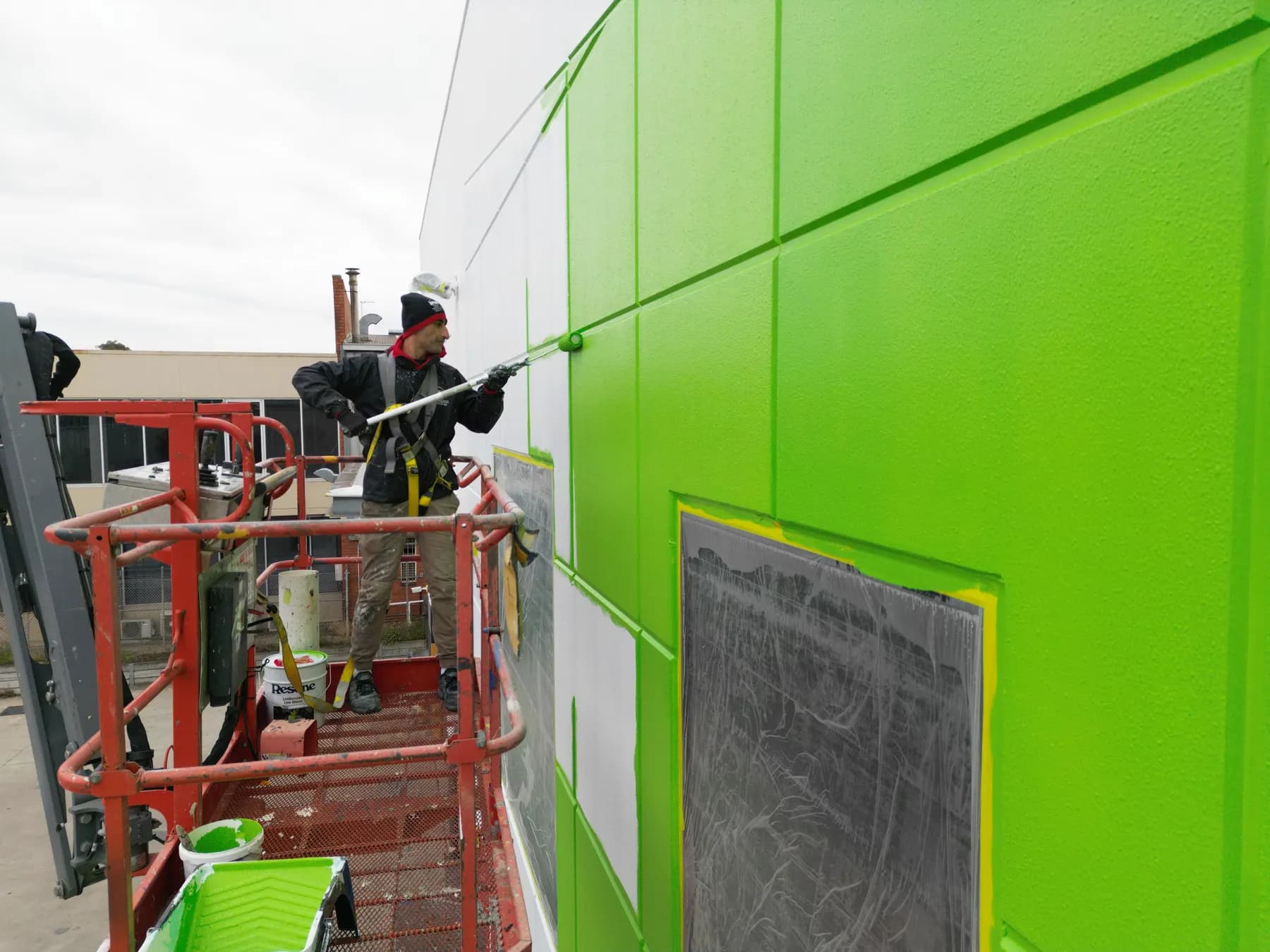 A harnessed painter on a scissor lift rolling bright green onto a panelled factory wall with an extension pole