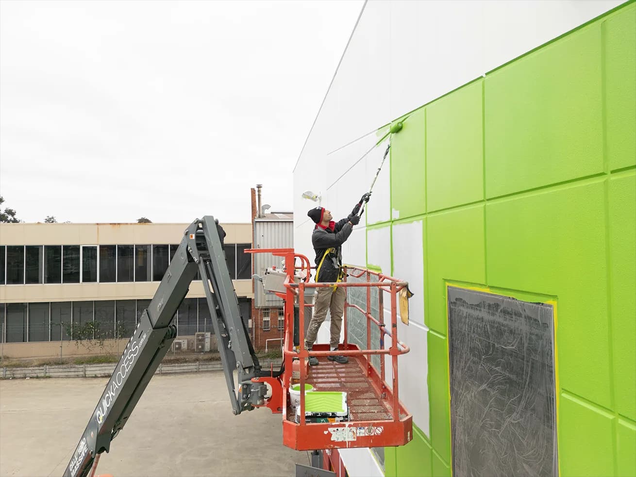 A painter working from a boom lift, harnessed, cutting in the line between white and green panels on a warehouse wall