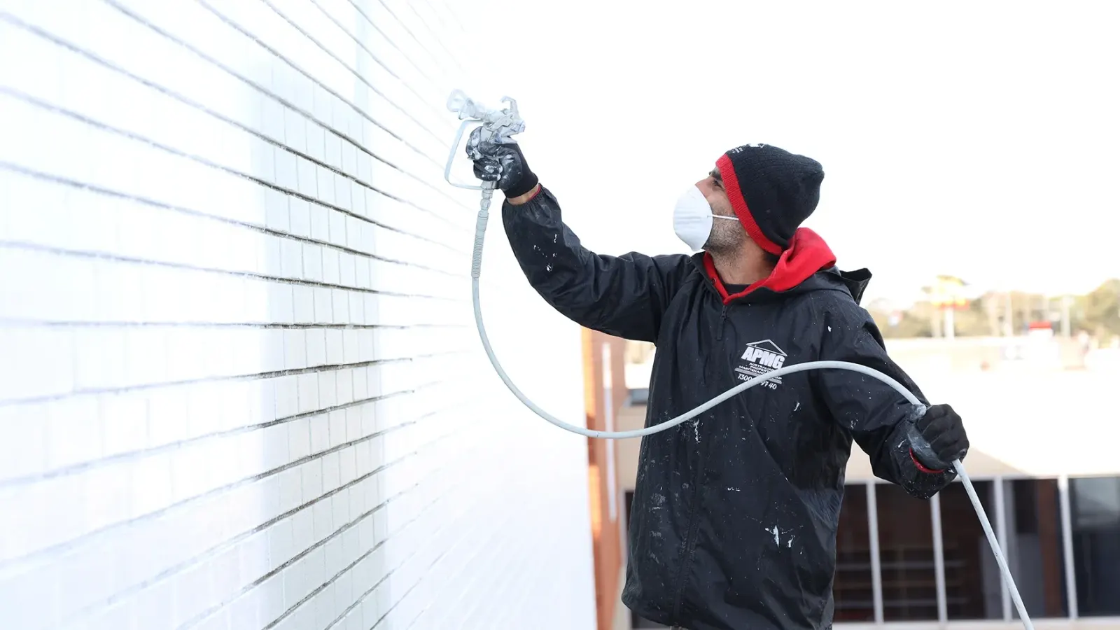 A painter in a respirator spraying the finish coat onto a face-brick elevation, spray line in hand