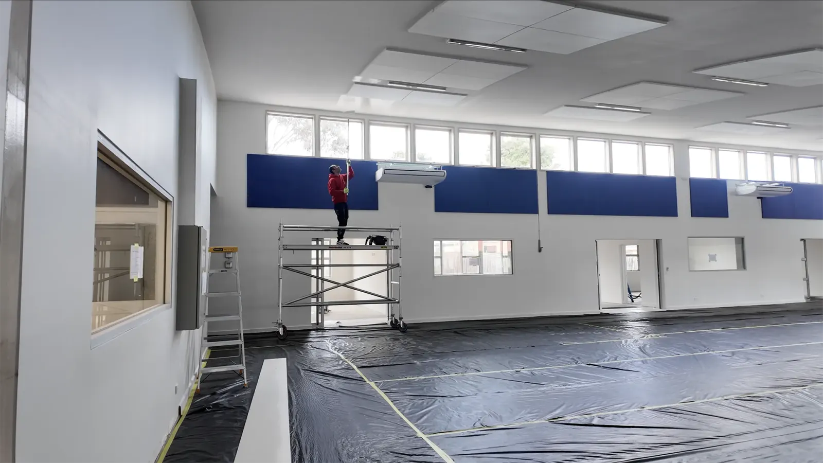 A painter on a mobile scaffold cutting in the blue band beneath the clerestory windows of a school hall, the whole floor sheeted in black plastic