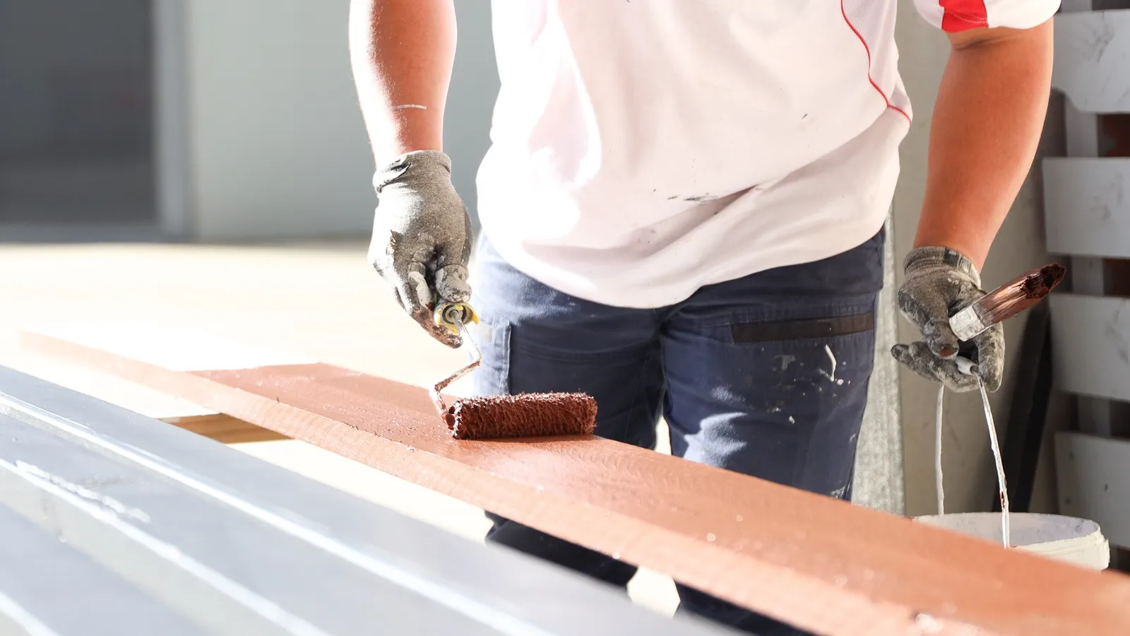 A painter rolling primer onto a length of trim laid across trestles on a site still under construction, the rest of the run stacked alongside