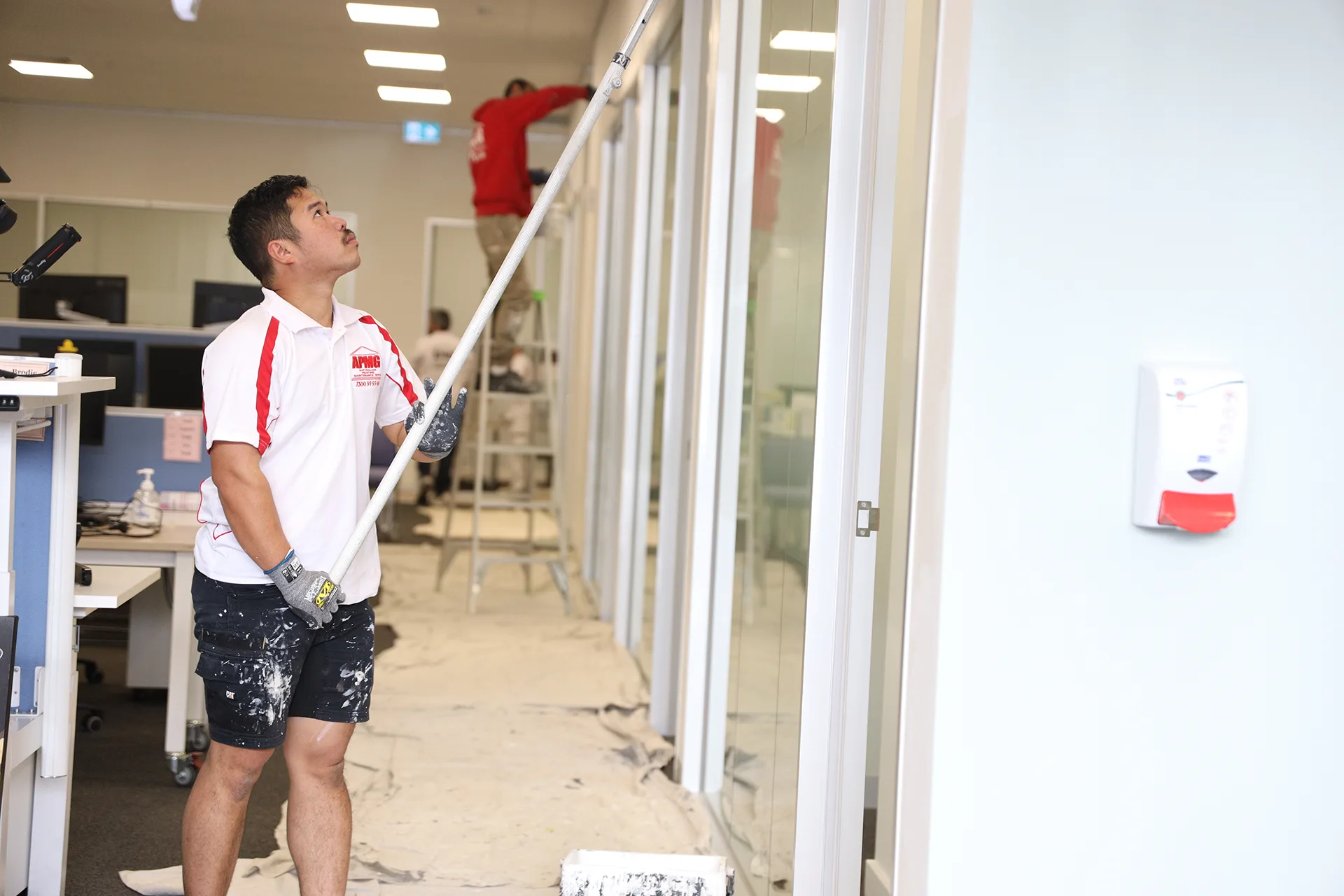 A painter running a pole roller down an office wall beside occupied desks and monitors, a second painter working the partition line behind